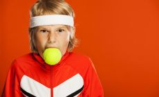 boy in tennis gear with tennis ball in his mouth and angry expression